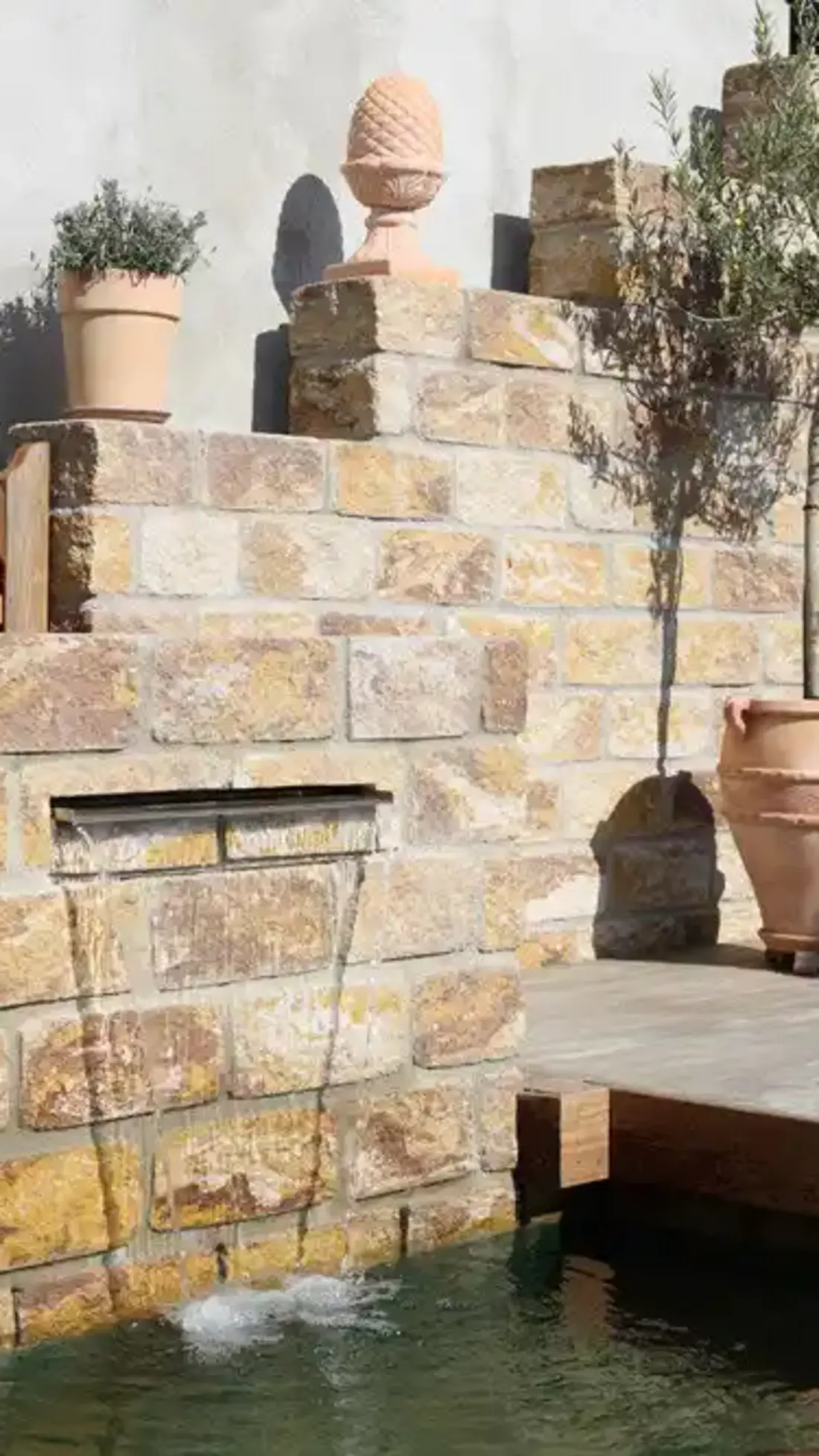 Mediterraner Garten mit Natursteinmauer wie auch integriertem Wasserauslauf und Holzdeckbelag